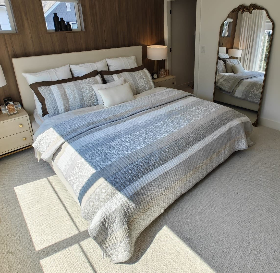 A modern bedroom featuring a bed dressed in blue and gray patterned bedding, set against a dark wood accent wall. The room includes a large arched mirror, bedside tables with lamps, and a light wood dresser in the foreground.